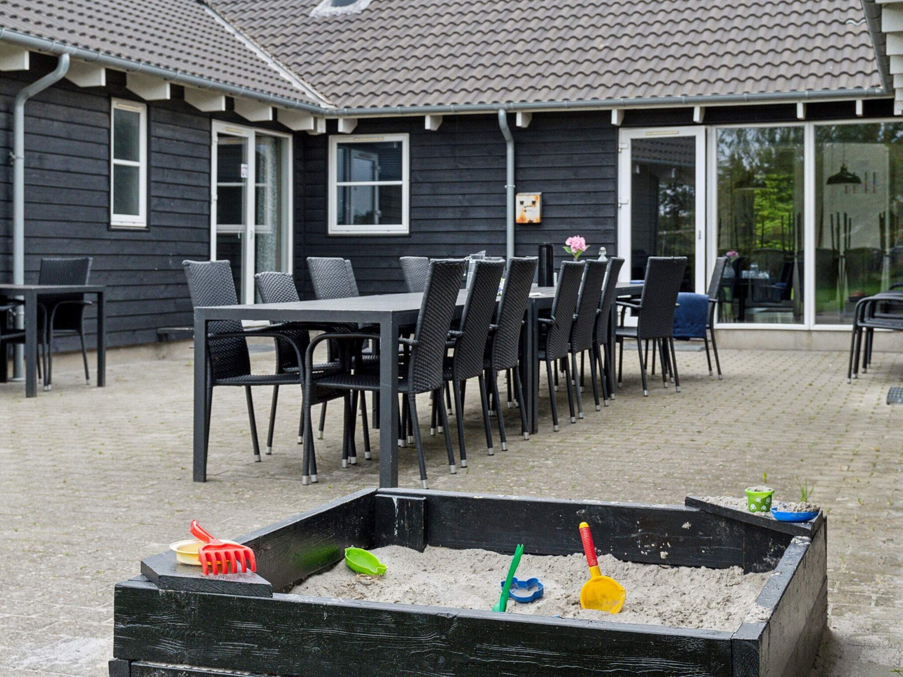 An outdoor patio with a long table and chairs, a sandbox with toys, and a building with windows and a door.