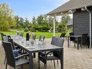 Une terrasse extérieure avec une longue table et des chaises, entourée de verdure et d’un bâtiment.
