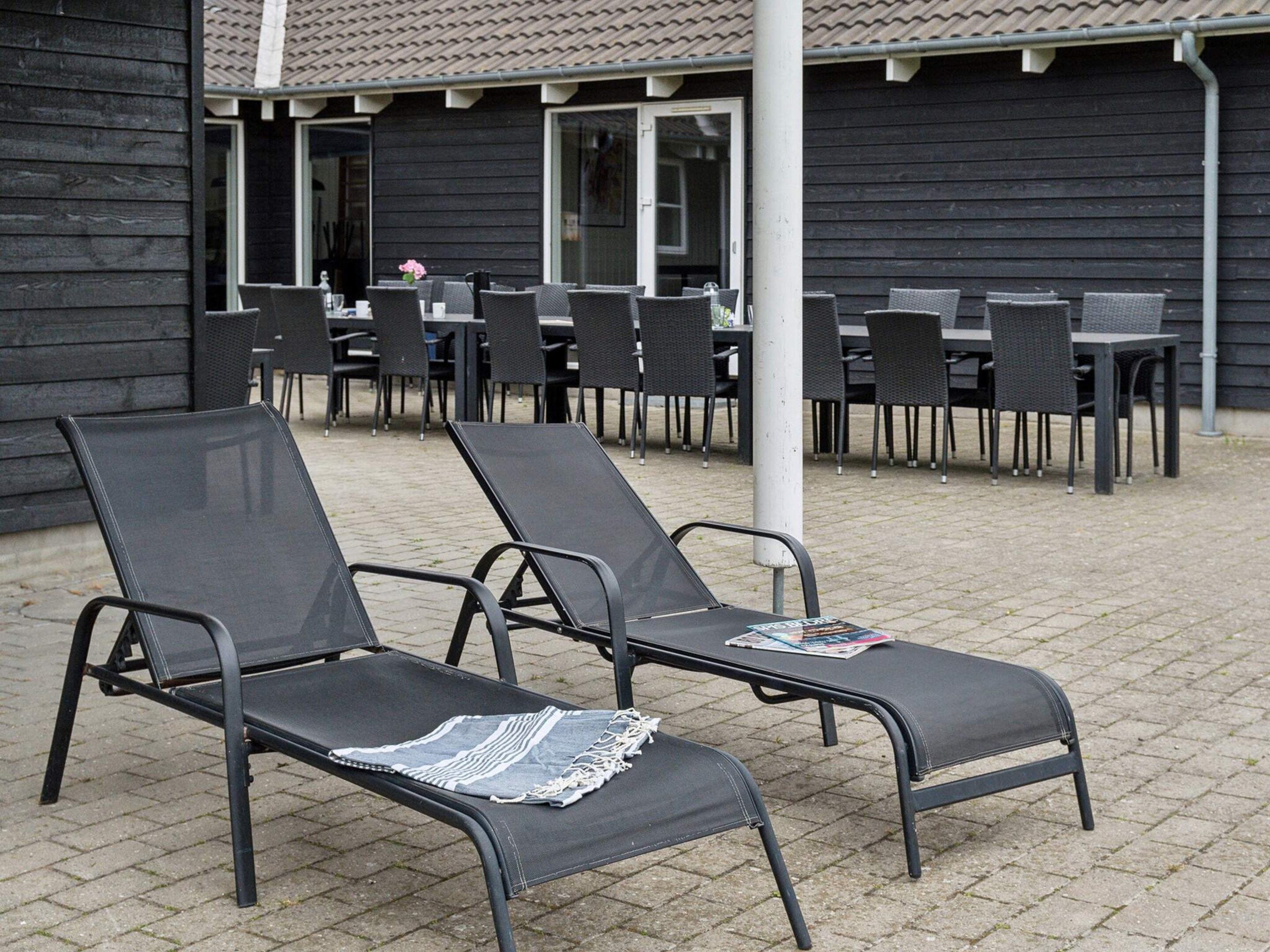 Deux fauteuils confortables, dont l’un est agrémenté d’un magazine, sont placés devant un bâtiment dont le toit est recouvert de tuiles. On aperçoit en arrière-plan plusieurs tables et chaises.