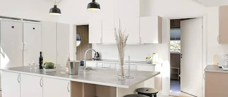 Una cocina moderna con armarios blancos, una isla central, dos taburetes de bar, luces colgantes y vistas al exterior.