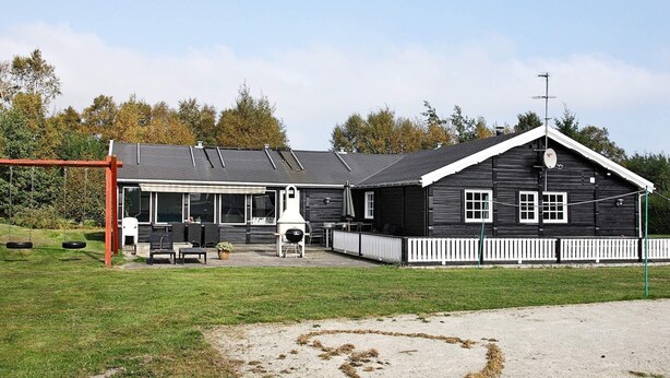 A black building with a white fence, a patio area with outdoor furniture, and a swing set. - 5 Star Holiday Home in Hals (Hals)