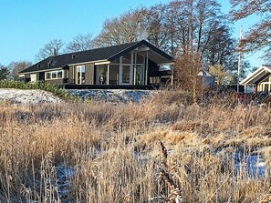 A modern house with a large windowed porch, surrounded by tall grasses and trees.