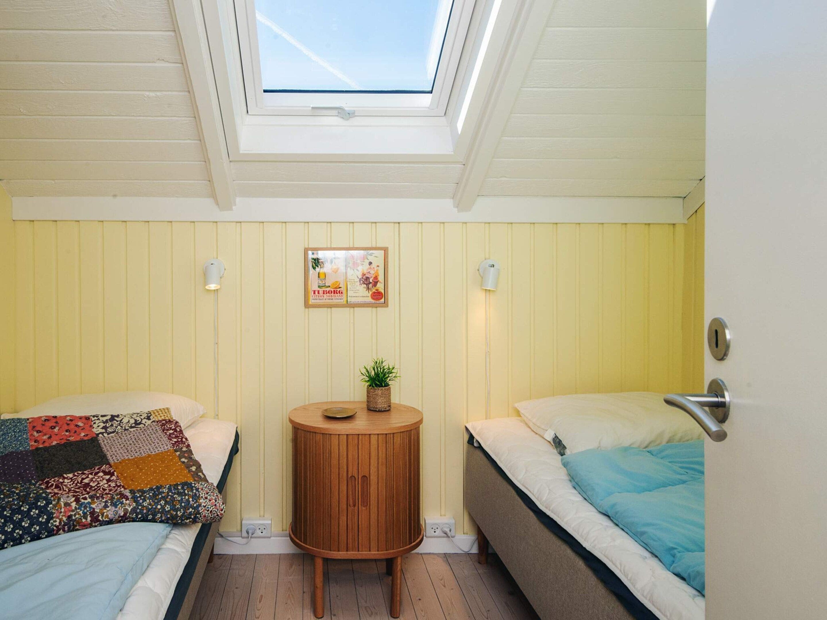 A room with two beds, a small round wooden table, a potted plant, a framed picture on the wall, and a skylight above.