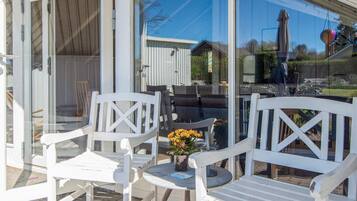 A patio area with white chairs and a table, a glass door, and a view of a house and garden.