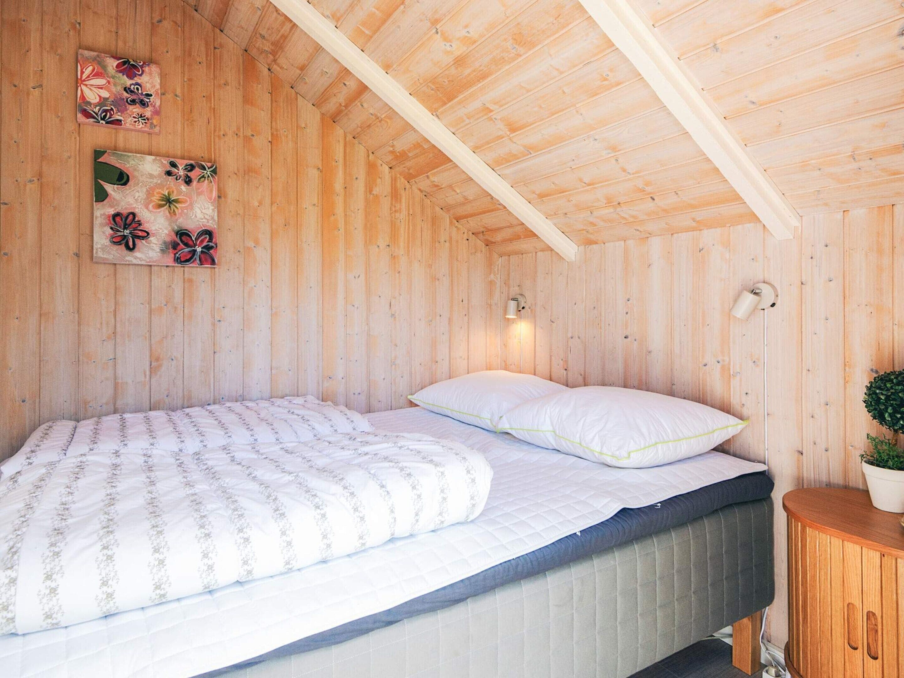 Un dormitorio de cabaña de madera con una cama, dos lámparas fijadas a la pared y dos cuadros con motivos florales.