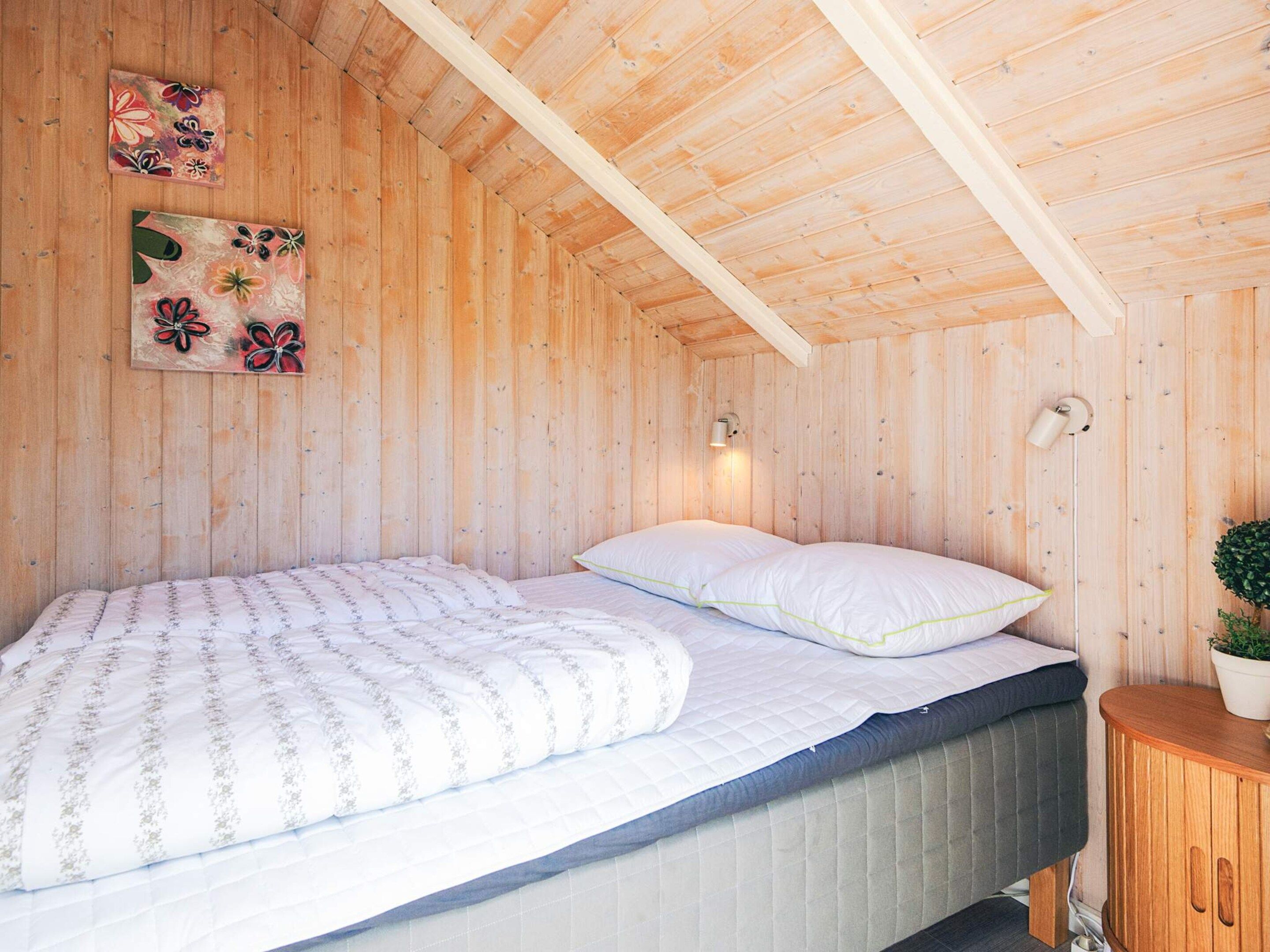 Una habitación de madera con una cama, una lámpara en la pared y dos cuadros con motivos florales.