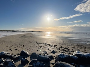 A beach with a clear sky, the sun low on the horizon, and large rocks in the foreground.