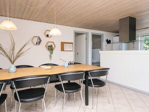 Uma área de refeições moderna com mesa de madeira e cadeiras pretas, uma decoração de parede hexagonal e uma cozinha visível através de uma porta aberta.