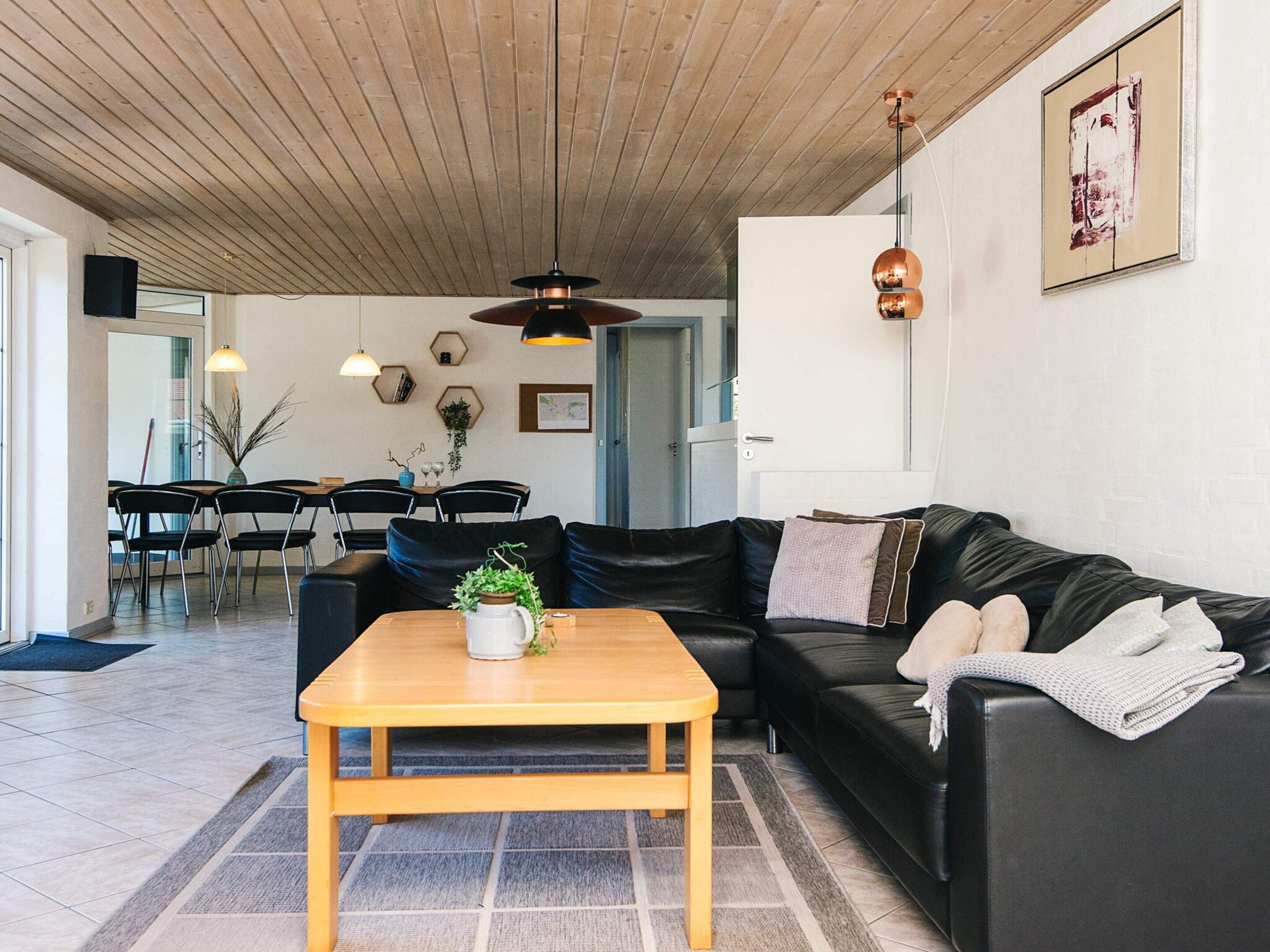 A modern living room with a wooden ceiling, a black leather sofa, a wooden coffee table, and a dining area with chairs and a table.