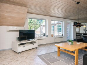 A modern living room with a television on a white cabinet, a wooden table, and a large window with a view of the outside. - 10 Person Holiday Home in Blavand (Blavand)
