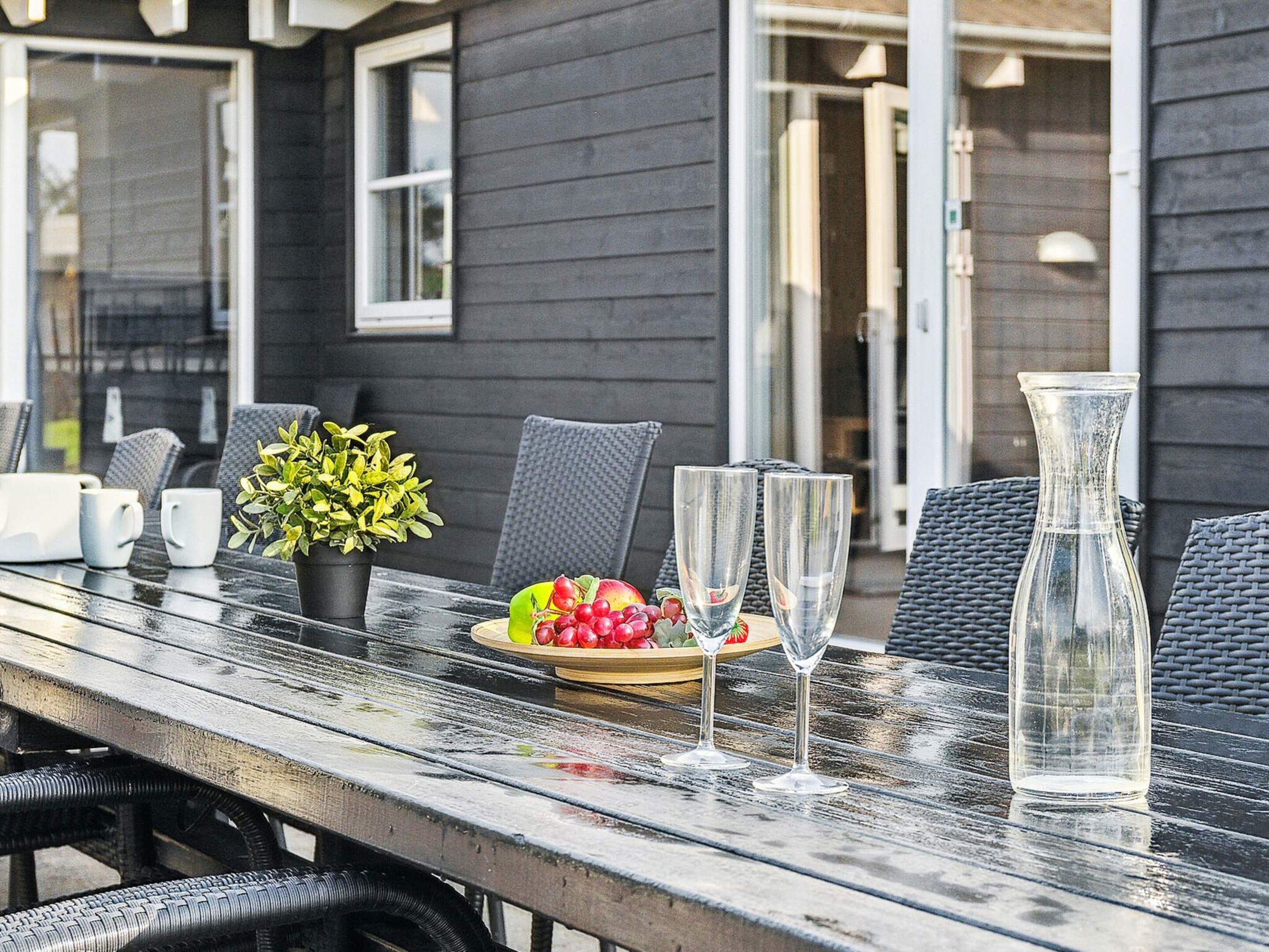 An outdoor dining area with a wooden table, chairs, a potted plant, a fruit bowl, and wine glasses.