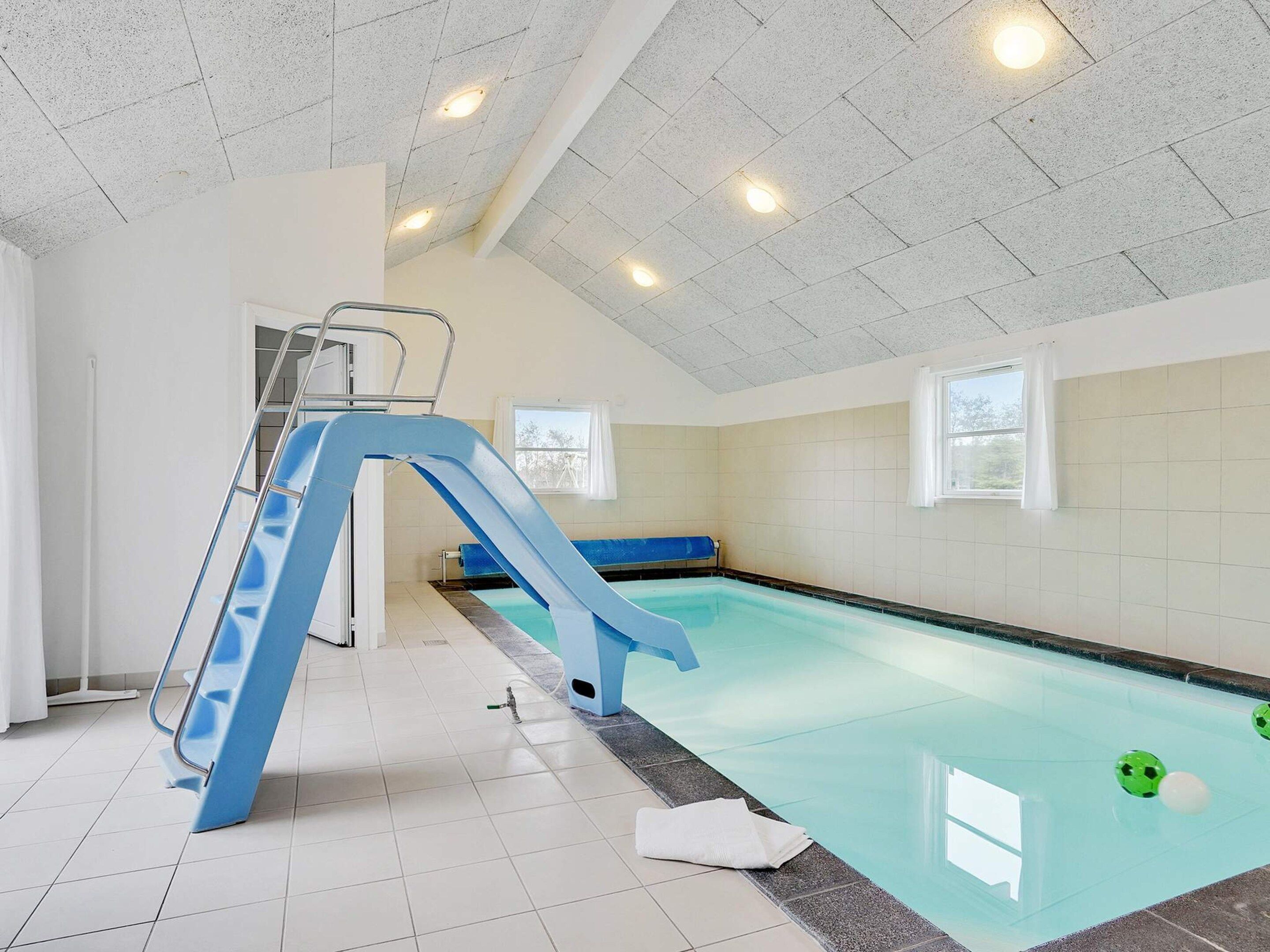 Una piscina interior amb un tobogan blau, una escala i un banc.