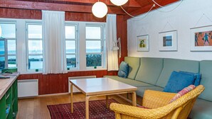 Een gezellige kamer met een houten plafond, een groene bank, een rieten stoel, een tafeltje en grote ramen met gordijnen.