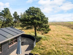 A building with a curved roof, a patio with a table and chairs, and a view of a grassy field with trees. - 5 Star Holiday Home in Blavand (Blavand)