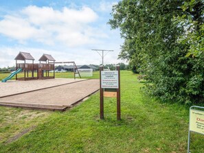Une aire de jeux avec un toboggan et des balançoires, entourée d’herbe et d’arbres.