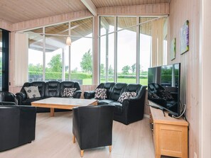 Un salon spacieux avec des canapés en cuir, une table basse en bois et un téléviseur posé sur un meuble en bois. De grandes fenêtres offrent une vue sur l’extérieur.