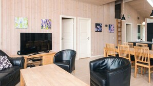 A living room with a TV, black leather sofa, wooden coffee table, and a dining area with wooden chairs and a table. - 5 Star Holiday Home in Ansager (Ansager)