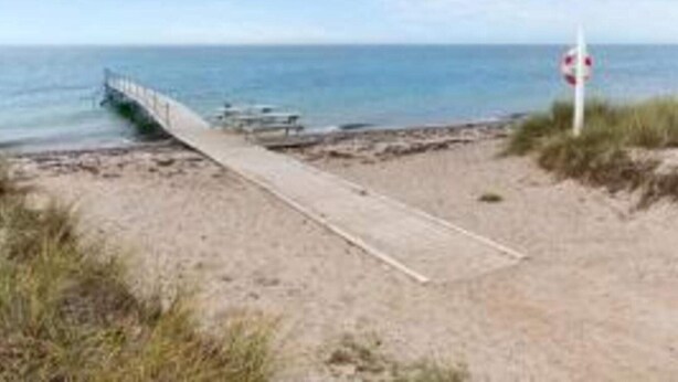Ein Strand mit einer Holzpromenade, die zum Wasser führt, einem Rettungsschwimmer-Schild und einem wolkenlosen Himmel.