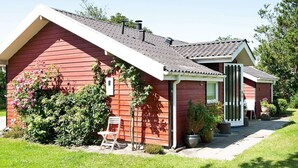 A red single-story house with a sloped roof, a front porch, and a small garden area with a chair and potted plants.