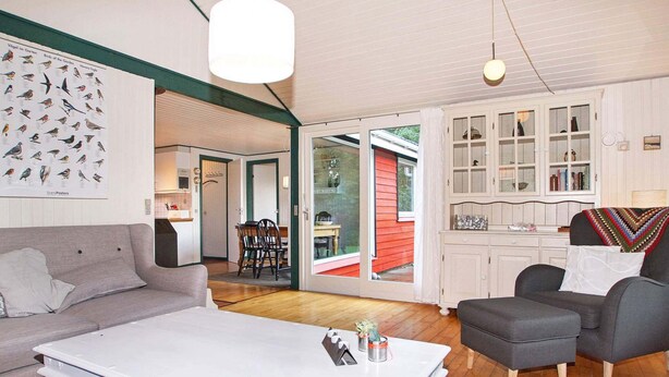 A spacious living room with a white coffee table, grey sofas, and a dining area with a red exterior visible through glass doors.