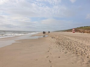 A wide sandy beach with footprints leading to the water, a red and white lifeguard tower, and a grassy hill in the background. - 5 Star Holiday Home in Ringkobing (Ringkobing)