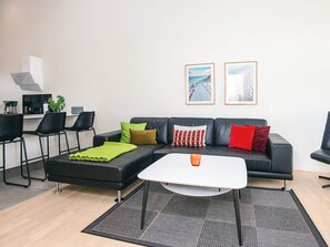 Un salon moderne avec un canapé en cuir noir, une table basse blanche et un coin cuisine à l’arrière-plan.
