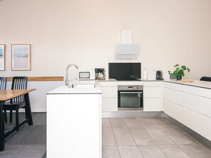 A modern kitchen with white cabinetry, a built-in oven, a microwave, and a sink. There is a dining table with chairs, a potted plant, and framed pictures on the wall. - 5 Star Holiday Home in Ringkobing (Ringkobing)