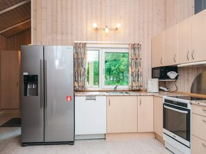 A modern kitchen with a stainless steel refrigerator, a built-in dishwasher, a window with curtains, wooden cabinets, and a microwave. - 4 Star Holiday Home in Ansager (Ansager)