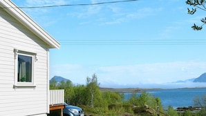 A white house with a car parked in front, a grassy area, and a lake with mountains in the background. - Holiday Home in Leknes (Vestvågøy)