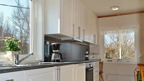 A modern kitchen with white cabinets, a built-in oven, a coffee maker, and a window with a view of trees. - Holiday Home in Leknes (Vestvågøy)