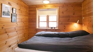 A cozy cabin room with wooden walls, a bed with two pillows, a window with a view, and a wall-mounted lamp. - Seaside Escape in Ho - By Traum Ferienwohnungen (Blavand)