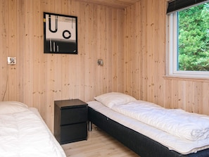 A room with wooden walls, two single beds, a small bedside table, and a window with a view of greenery.