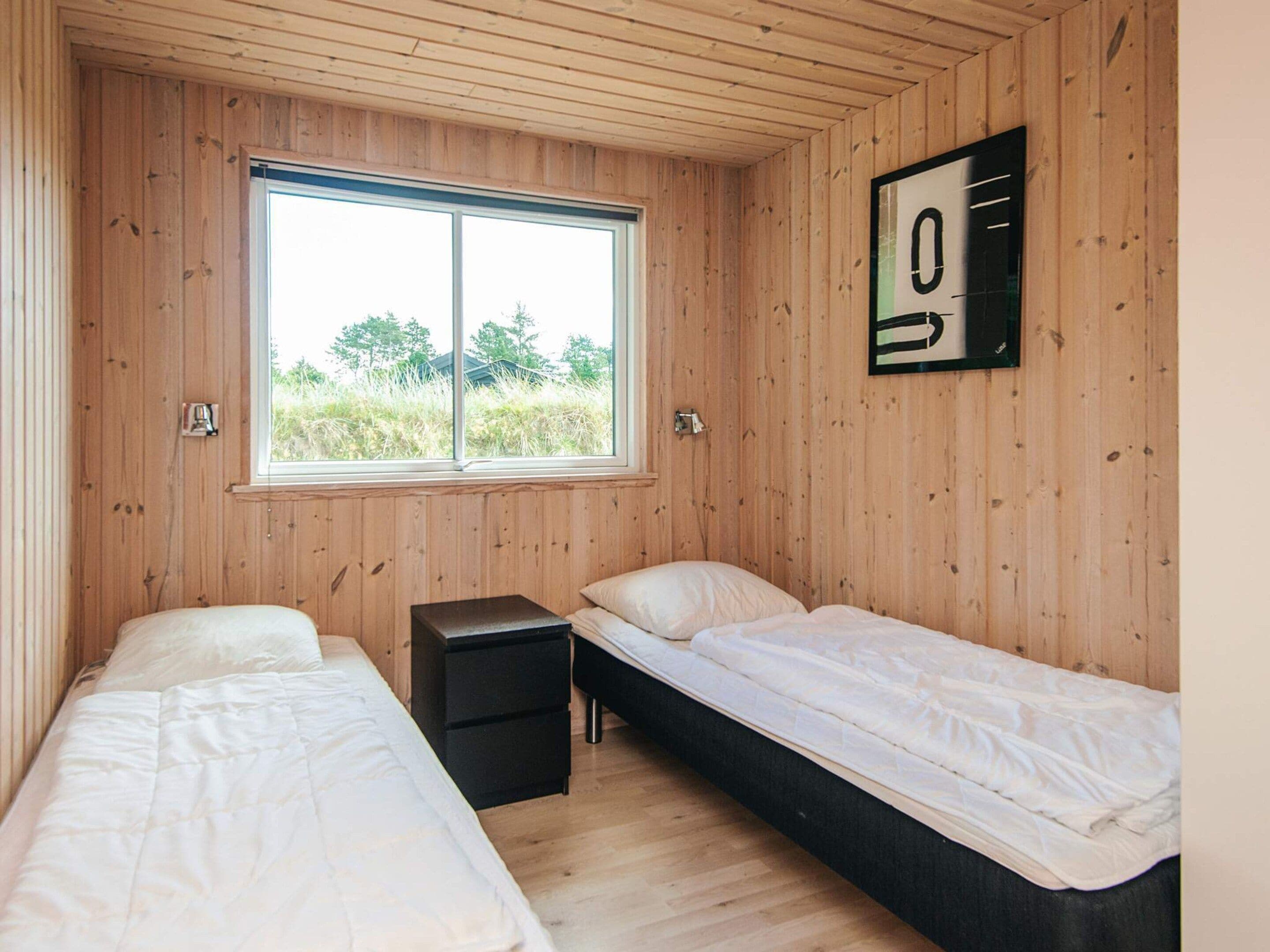 Ein Zimmer mit zwei Einzelbetten, einem kleinen schwarzen Nachttisch, einem Fenster mit Blick ins Grüne und einem an der Wand befestigten Nummernschild.