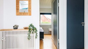 A hallway with a white radiator, a wooden console table with decorative items, a dark door, and a view into a room with a bed and a window. - 4 Star Holiday Home in Ulfborg (Ulfborg)