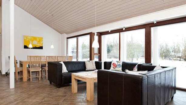 Un salon moderne avec un canapé d’angle, une table à manger avec des chaises et de grandes fenêtres donnant sur l’extérieur.