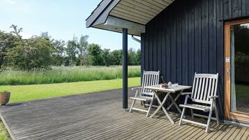 A wooden deck with two white chairs and a table, a dark wooden building with a glass door, and a grassy field in the background.