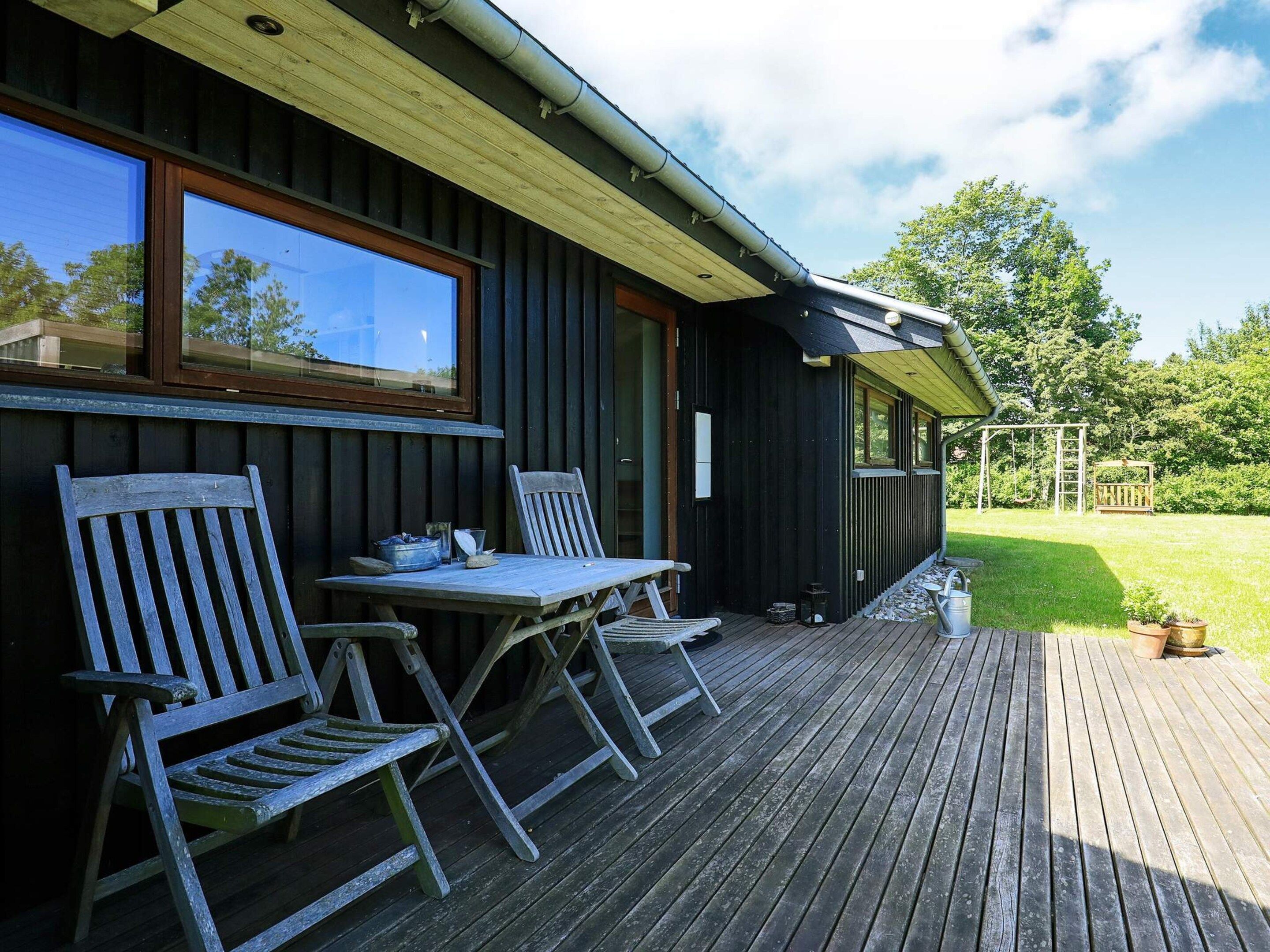 A wooden deck with two chairs and a table, a building with dark siding and a window, and a grassy yard with trees.