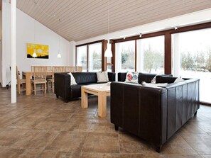 A modern living room with a sectional sofa, a wooden coffee table, and a dining area with chairs and a table. Large windows provide natural light and views of the outdoors. - 4 Star Holiday Home in Dannemare (Dannemare)