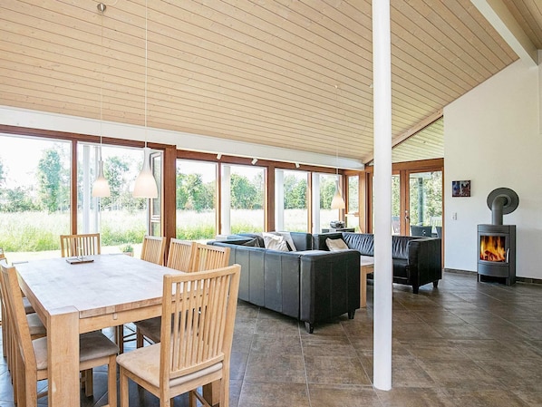A spacious indoor area with a wooden ceiling, large windows, a dining table with chairs, a sofa, and a wood-burning stove.