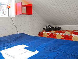 A room with a sloped ceiling, a bed with a patterned bedspread, a red storage unit, and a window with a view of a building.