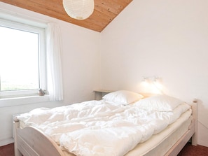 A bedroom with a wooden ceiling, a window with white curtains, a bed with white bedding, and a wall-mounted light fixture.