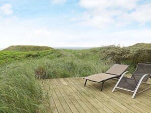 A wooden deck with two sun loungers, overlooking a grassy landscape with dunes and a view of the sea.