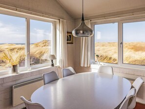 A room with a large round table, white chairs, a hanging light fixture, and large windows with a view of a field. - 4 Star Holiday Home in Lokken (Lokken)