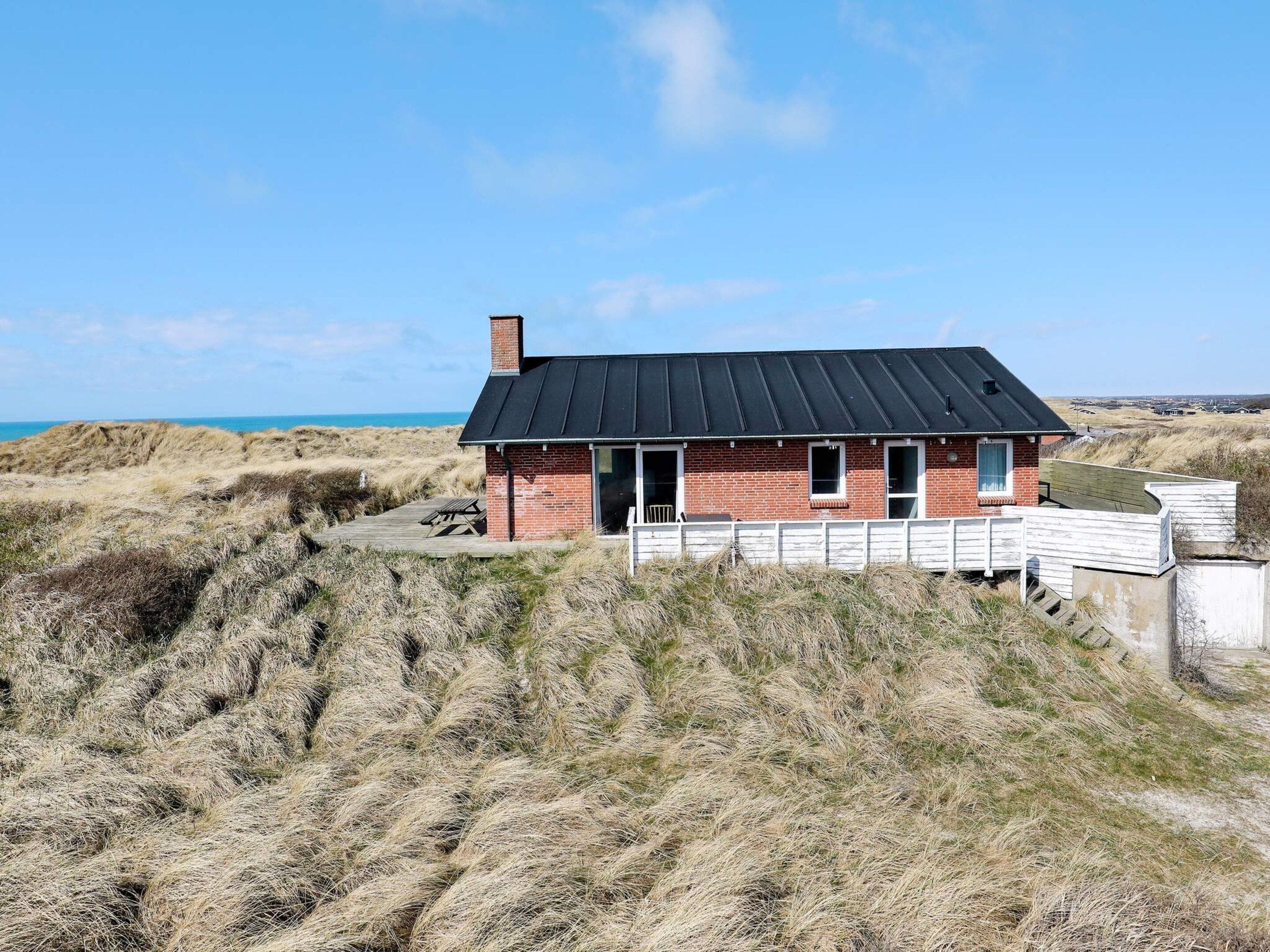 A single-story brick house with a black roof, a wooden deck, and a white fence.