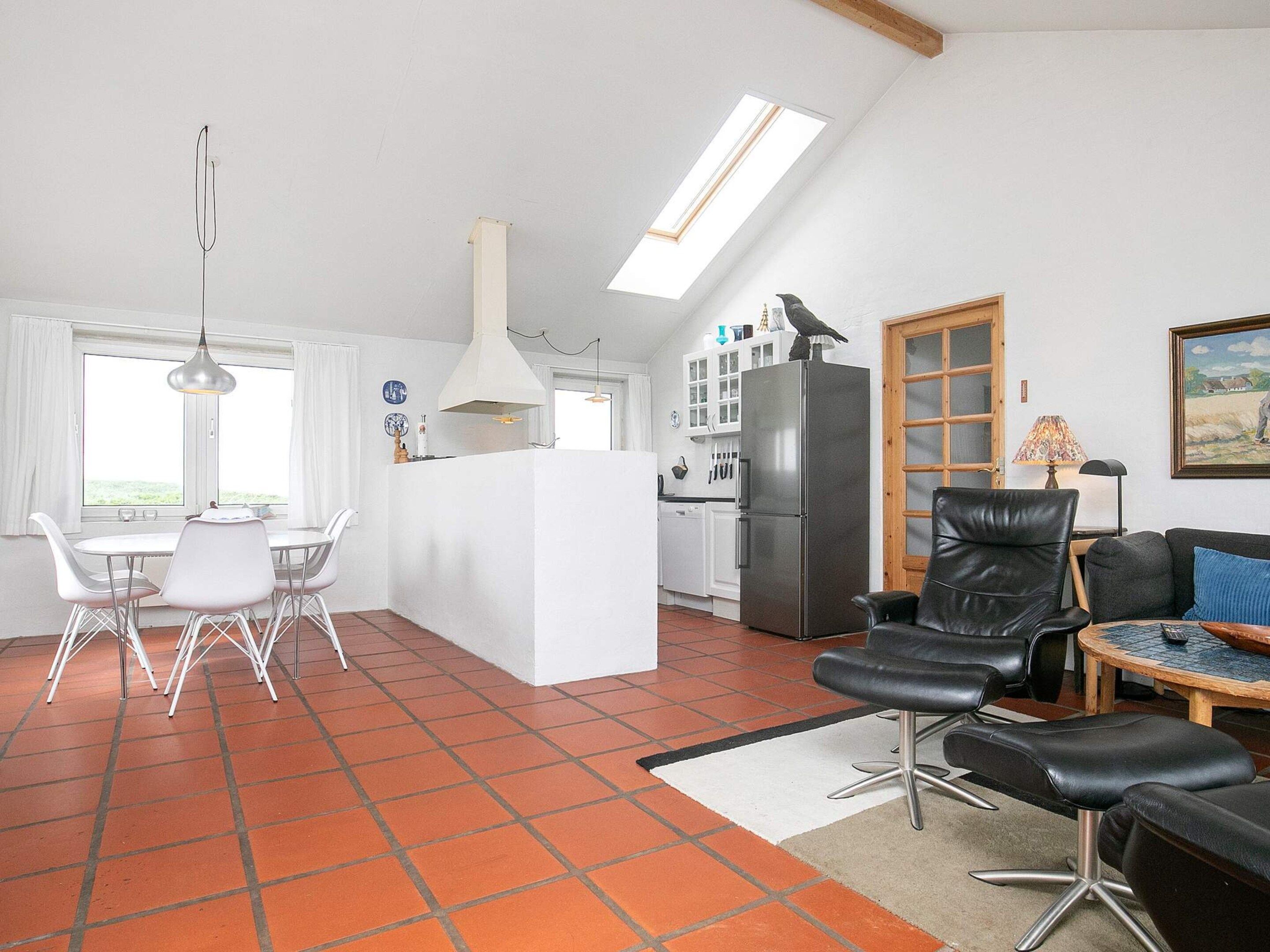 A modern kitchen with a central island, a dining area with white chairs, a black leather sofa, and a tiled floor.