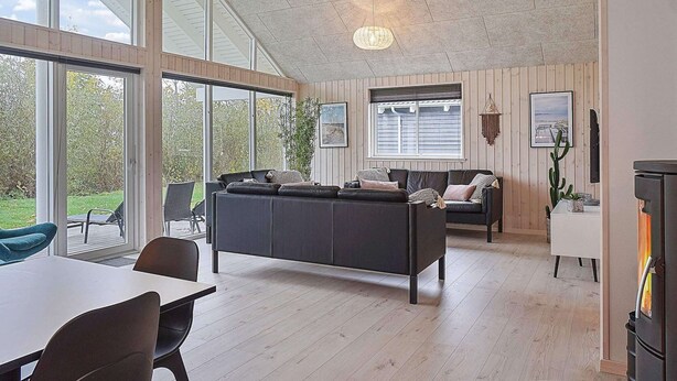 A modern living room with a large black sofa, a dining table with chairs, a fireplace, and large windows offering a view of the outdoors. - Holiday Home in Kappeln (Kappeln)