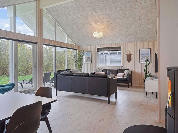 Ein modernes Wohnzimmer mit einer großen schwarzen Couch, einem Esstisch mit Stühlen, einem Kamin und großen Fenstern mit Blick ins Freie.