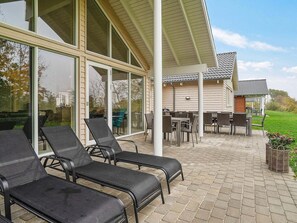 A covered outdoor seating area with chairs and tables, large windows, and a view of a house in the background. - Holiday Home in Kappeln (Kappeln)