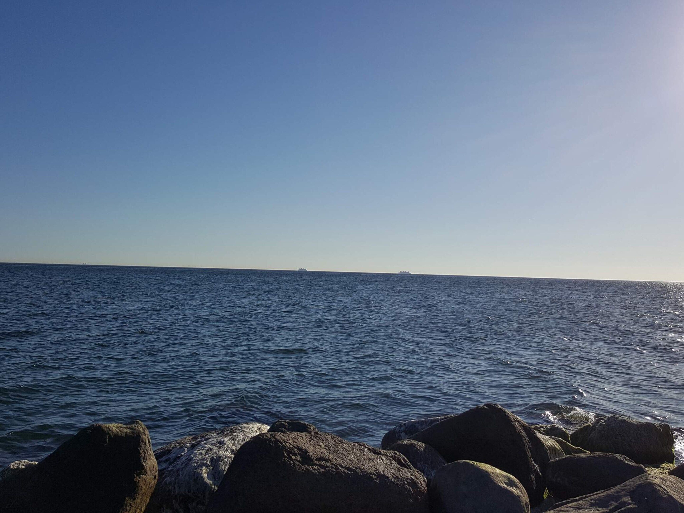 A clear view of the sea with a distant ship on the horizon and large rocks in the foreground.