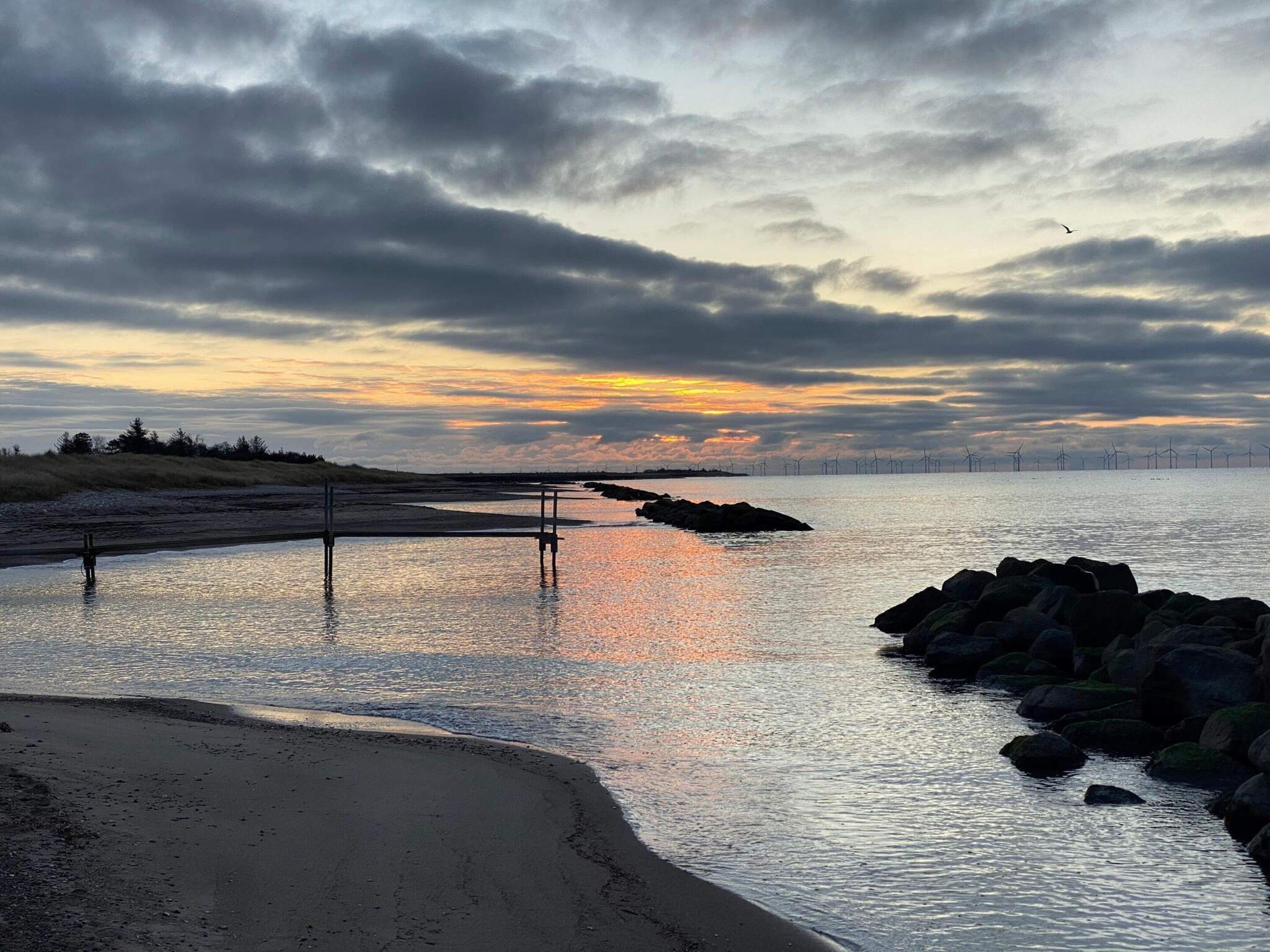 A beach with calm water, a rocky shoreline, and wind turbines in the distance.